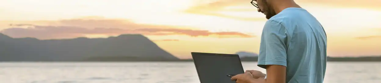 A person works on a mobile phone and laptop by a body of water, with cliffs in the distance. It is sunset.