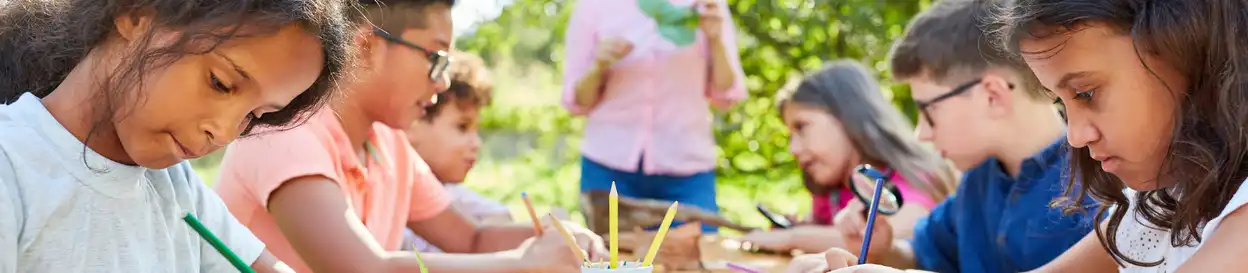 A summer camp - three young children are in frame working on a craft activity. A teacher's torso is seen at the back of the frame