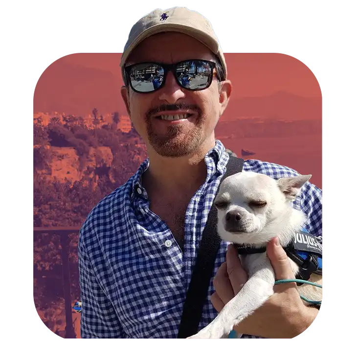 A man in sunglasses and a baseball cap smiles while holding a small white dog, with a scenic European landscape in