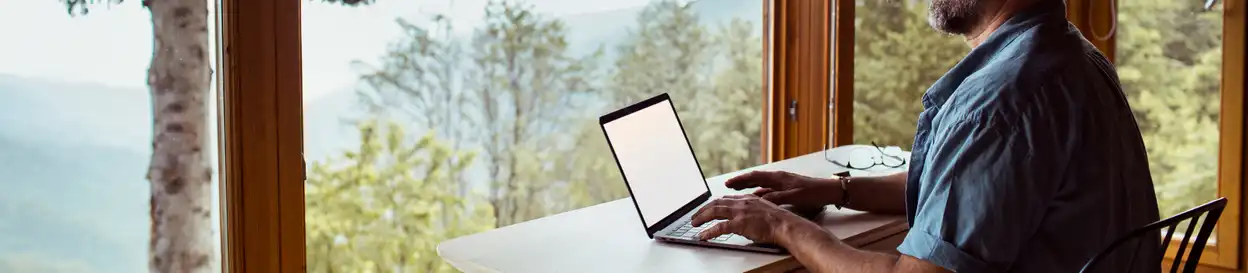 A man working on a laptop in front of a window with a scenic view