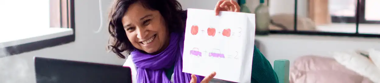 A female teacher holding up a piece of paper in front of a laptop with drawings on it