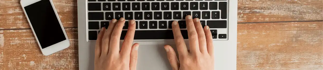 A person typing on a laptop with their phone to the left