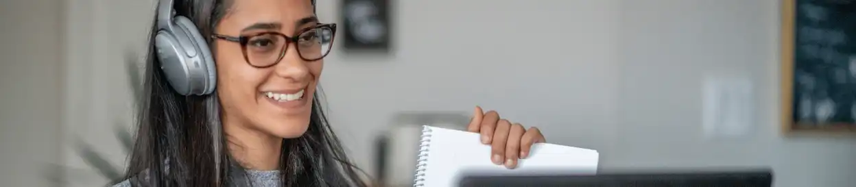 A woman with glasses wearing a headset and holding up a booklet in front of a laptop