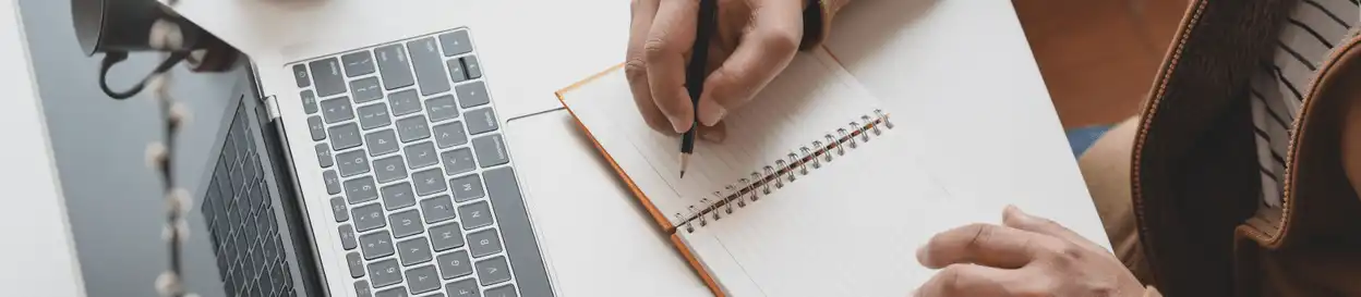 A person writing in a notebook while sitting in front of a laptop